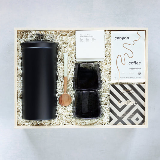 Gift set with black mugs, a wooden spoon, a black French Press, chocolate bar, candle, and coffee beans packed in a wooden box on a light gray background