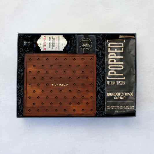 a poker gift set including the iron and glory dead man's hand poker chip and card set, bittermilk old fashioned cocktail mixer, popped bourbon espresso caramel popcorn and sugarfina caramels grouped together and packed in a standard gray carnaby & vine gift box and wrapped up in holiday lights on a light gray background.