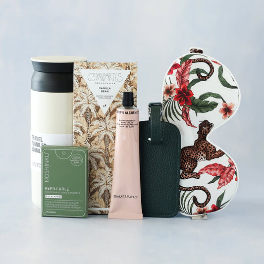 Collection of travel-friendly products including a tumbler, skincare items, a snack, and a luggage tag on a light gray background.