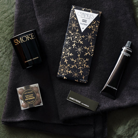 a holiday gift set that includes a black james perse cashmere scarf, compartes chocolate bar, sugarfina truffles, grown alchemist hand cream, and evermore london's smoke candle all laid out on a green linen fabric.