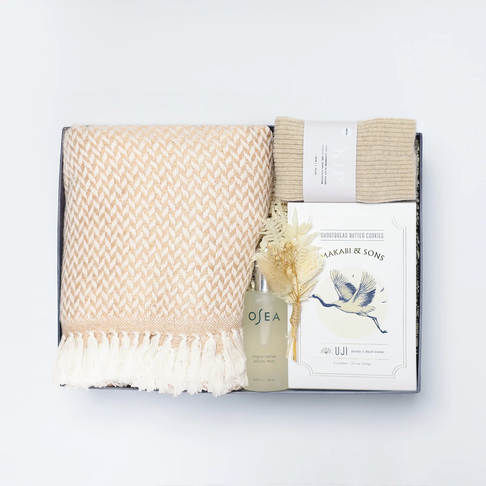 A gift set containing a beige cashmere throw, a bottle of OSEA Vagus Nerve Spray, and cookies by Makabi & Sons, all packaged in a box.