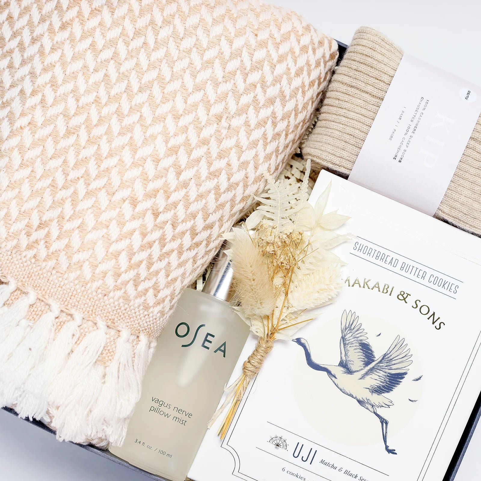 Close up of a beige blanket, socks, sleep spray, cookies, and dried flowers packaged in a gift box.