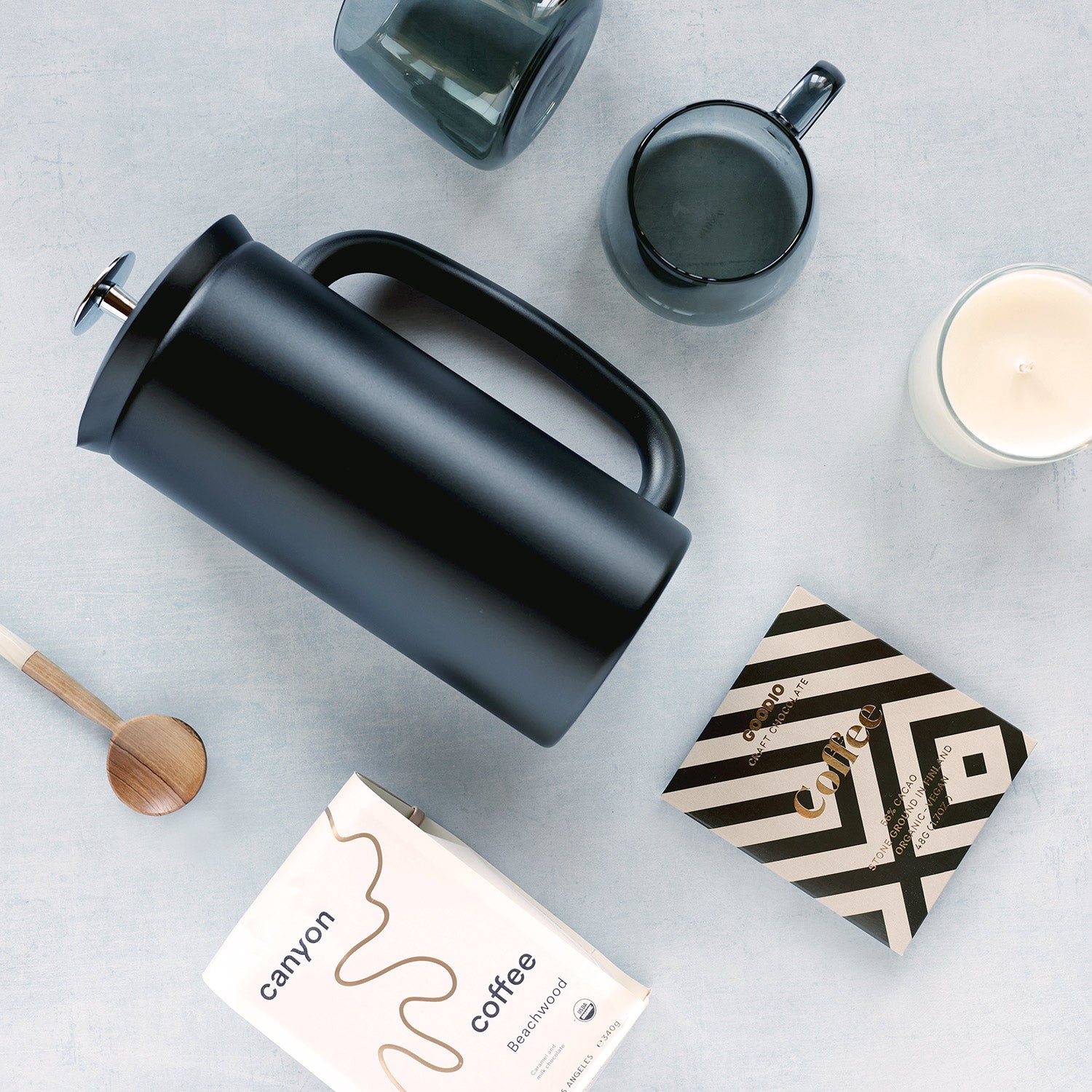 Black French press coffee maker on a light gray surface with a box of Canyon Coffee, smoke black mugs, and a candle.