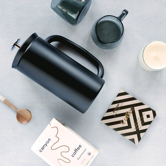 Black French press coffee maker on a light gray surface with a box of Canyon Coffee, smoke black mugs, and a candle.