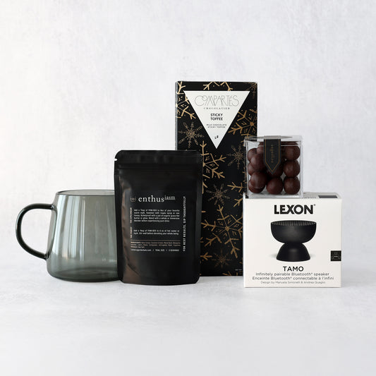 a holiday gift set including mushroom coffee, a lexon mini bluetooth speaker, sugarfina caramels, smoky gray mug and compartes sticky toffee chcolate bar grouped together on a light gray background.