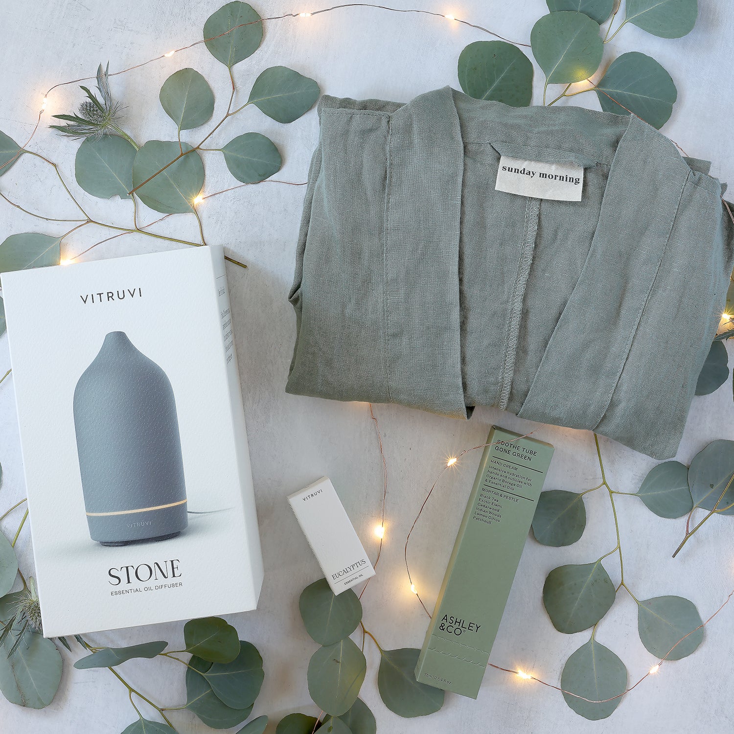 relaxing spa gift box with including sunday morning french linen robe, ashley and co hand cream, vitruvi diffuser and oil, grouped together with fresh greenery and twinkly lights on a light gray background.