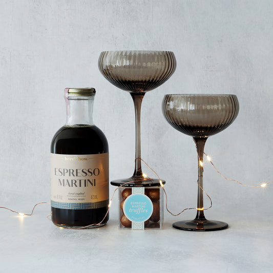 Bottle of Espresso Martini mixer, two smokey brown glasses, and a small box of truffles on a light gray background.