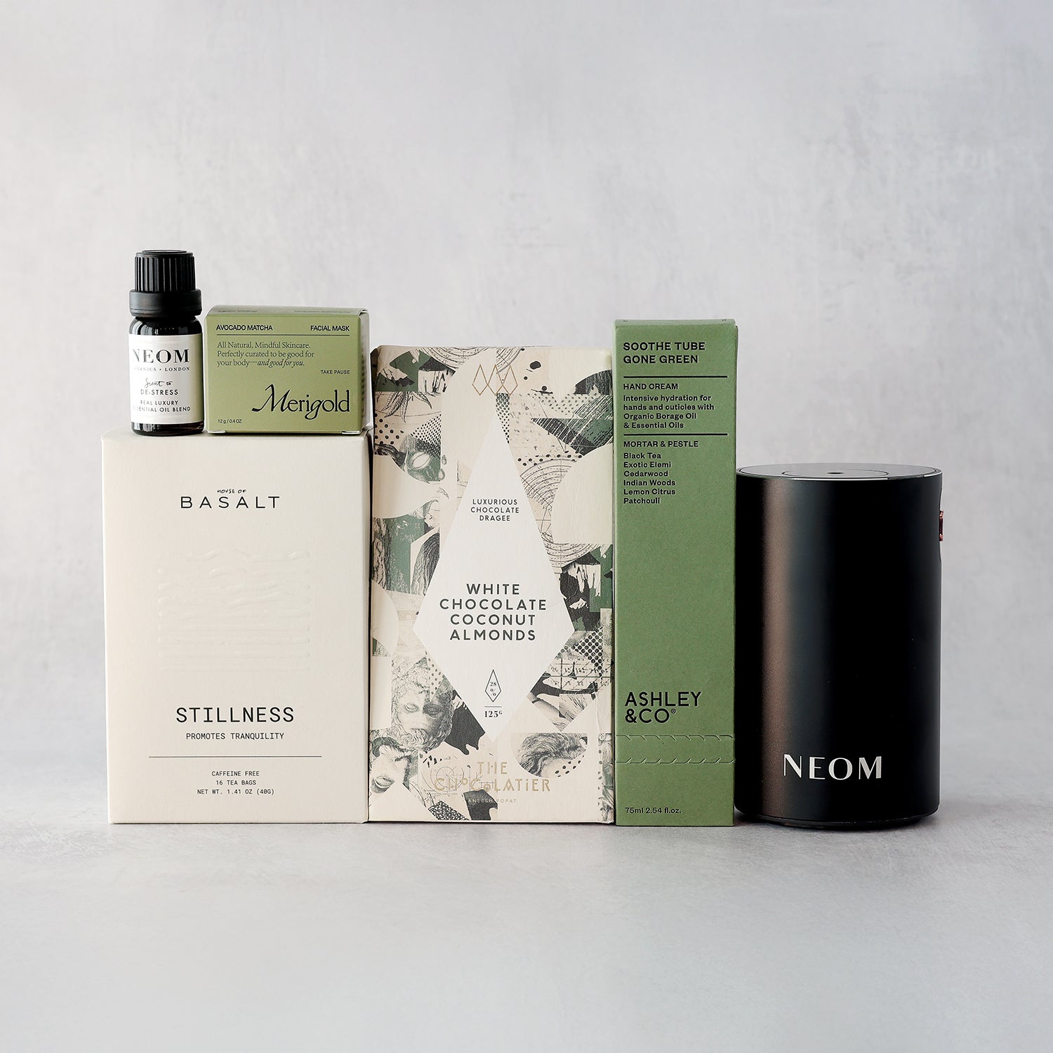spa gift box with green black and white elements including the neom portable diffuser, merigold avocado matcha face mask, ashley and co hand cream, basalt stillness tea, and the chocolatier white chocolate coconut almonds grouped together in front of a light background.