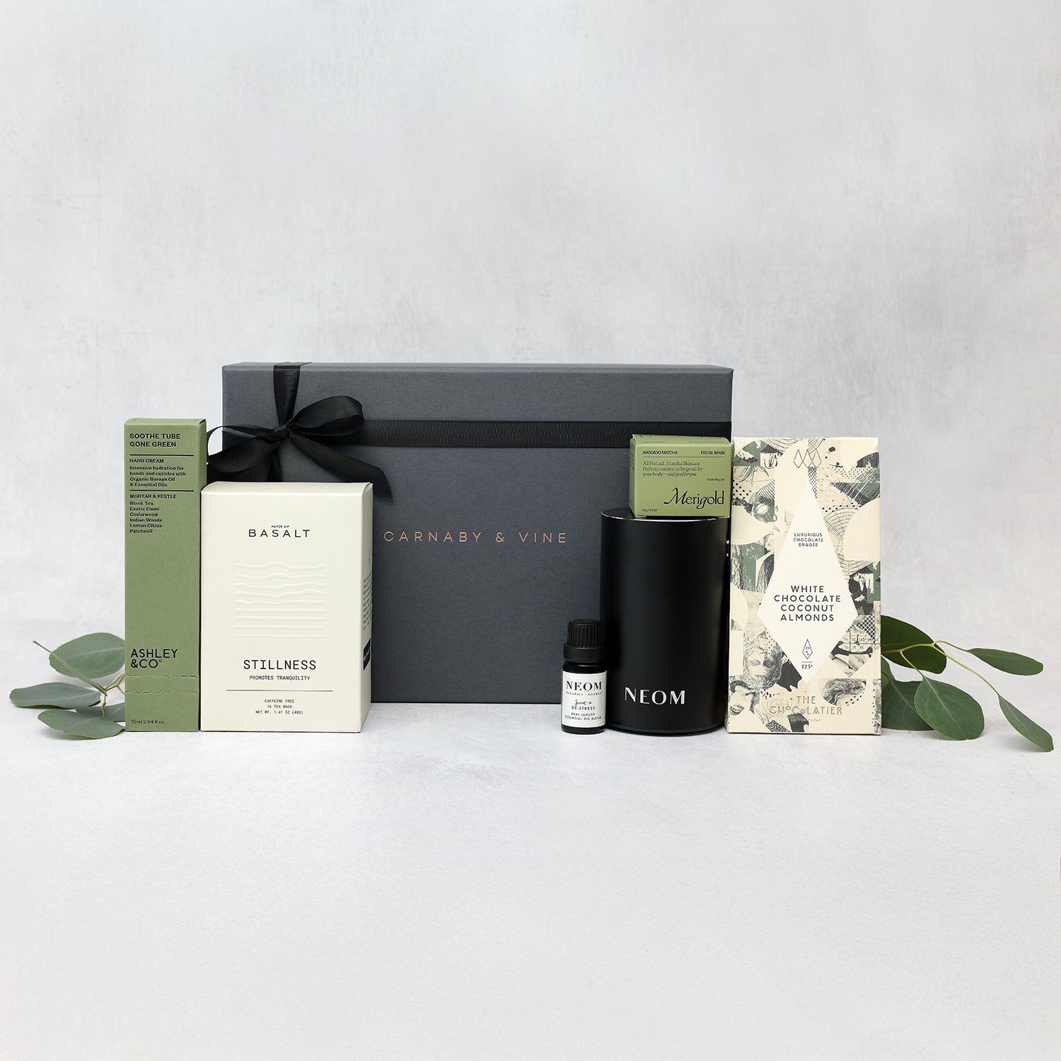 spa gift box with green black and white elements including the neom portable diffuser, merigold avocado matcha face mask, ashley and co hand cream, basalt stillness tea, and the chocolatier white chocolate coconut almonds grouped together in front of the carnaby and vine standard gift box on a light gray background.