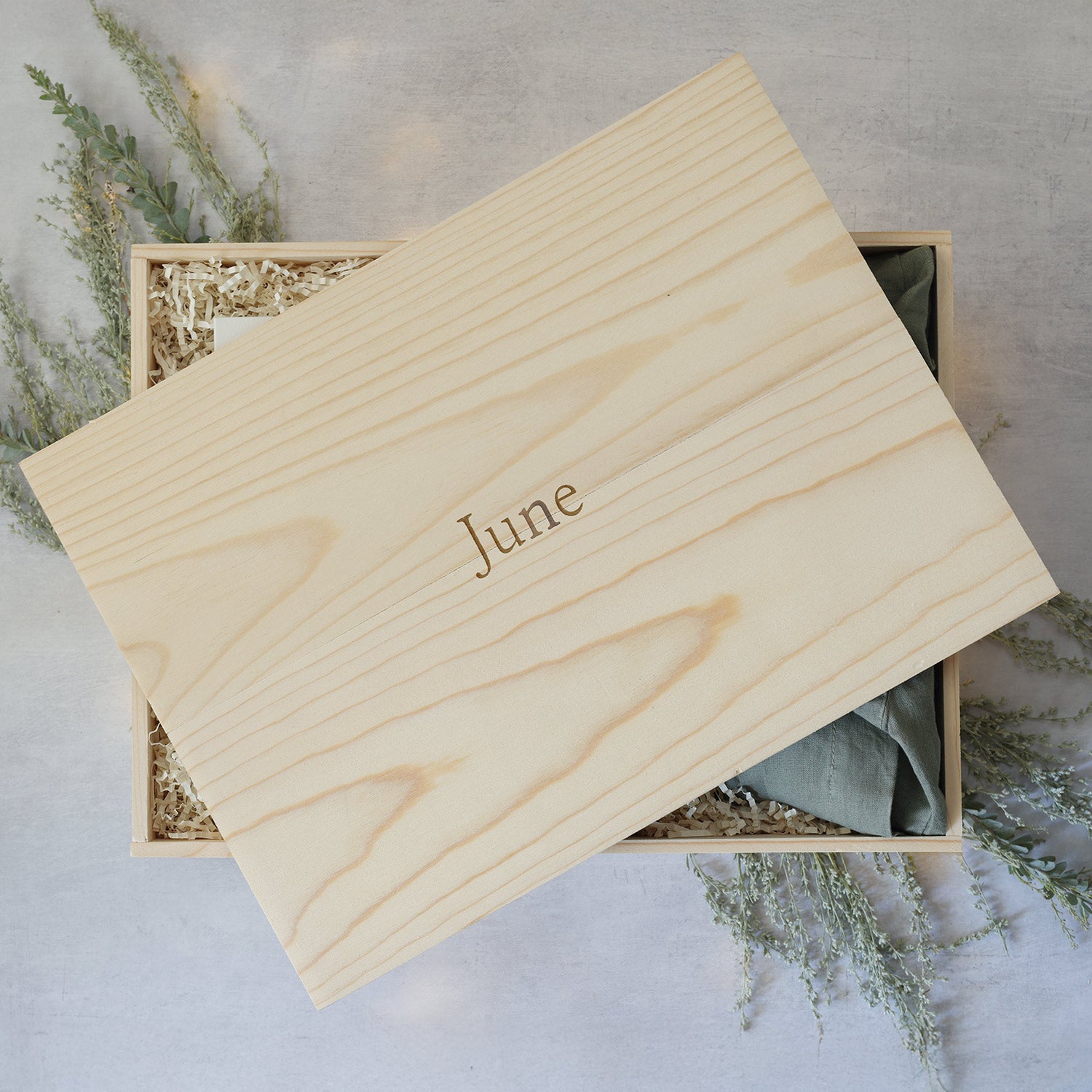 Wooden box with 'June' engraved on it, placed on a light gray surface with greenery.