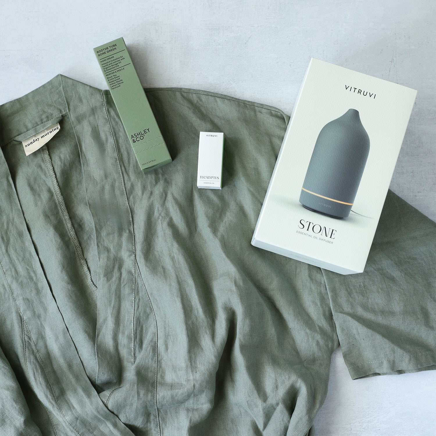 spa gifts box with eucalyptus essential oil and stone diffuser laid out on a green linen robe with a green hand lotion